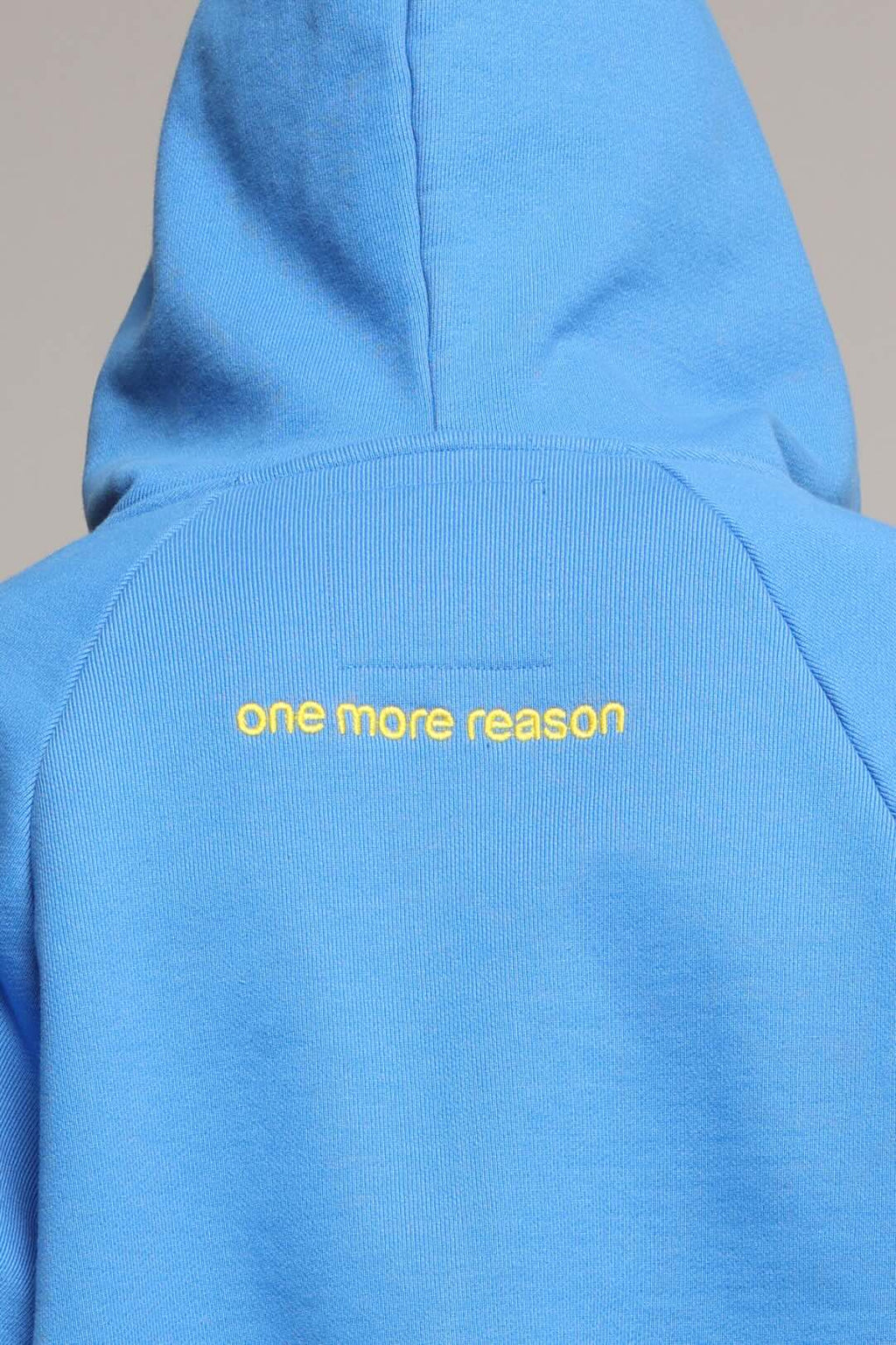 Close-up of the back of a light blue hoodie with yellow embroidered text reading 'one more reason' across the upper back from One More Reason, the European Premium Sportswear Brand.