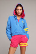 A woman wearing a bright blue zip-up hoodie with a pink-lined hood, yellow stripes on the sleeves, and matching hot pink sweat shorts with yellow stripes on the hem. All from One More Reason, the European Premium Sportswear Brand.