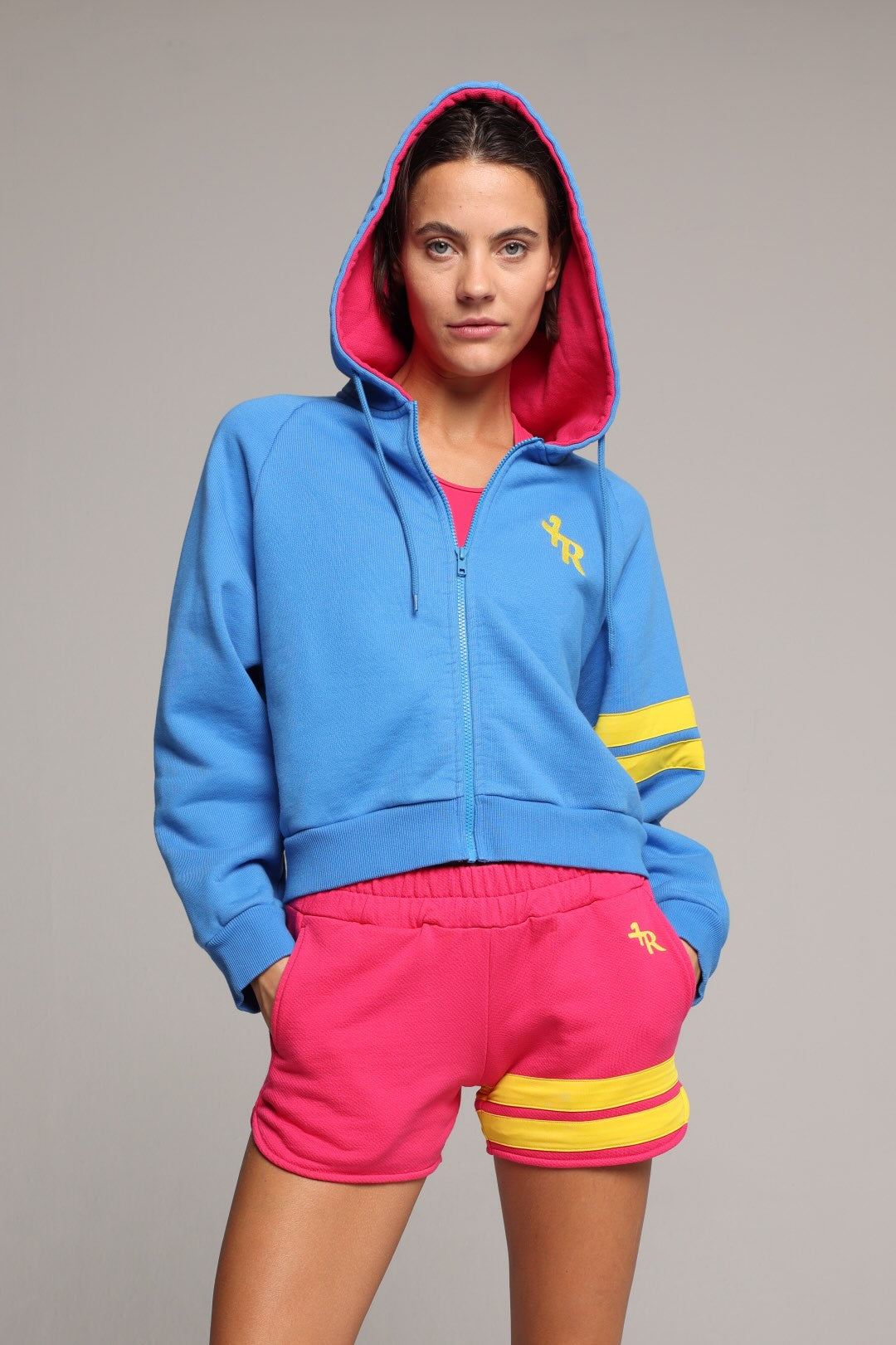 A woman wearing a bright blue zip-up hoodie with a pink-lined hood, yellow stripes on the sleeves, and matching hot pink sweat shorts with yellow stripes on the hem. All from One More Reason, the European Premium Sportswear Brand.