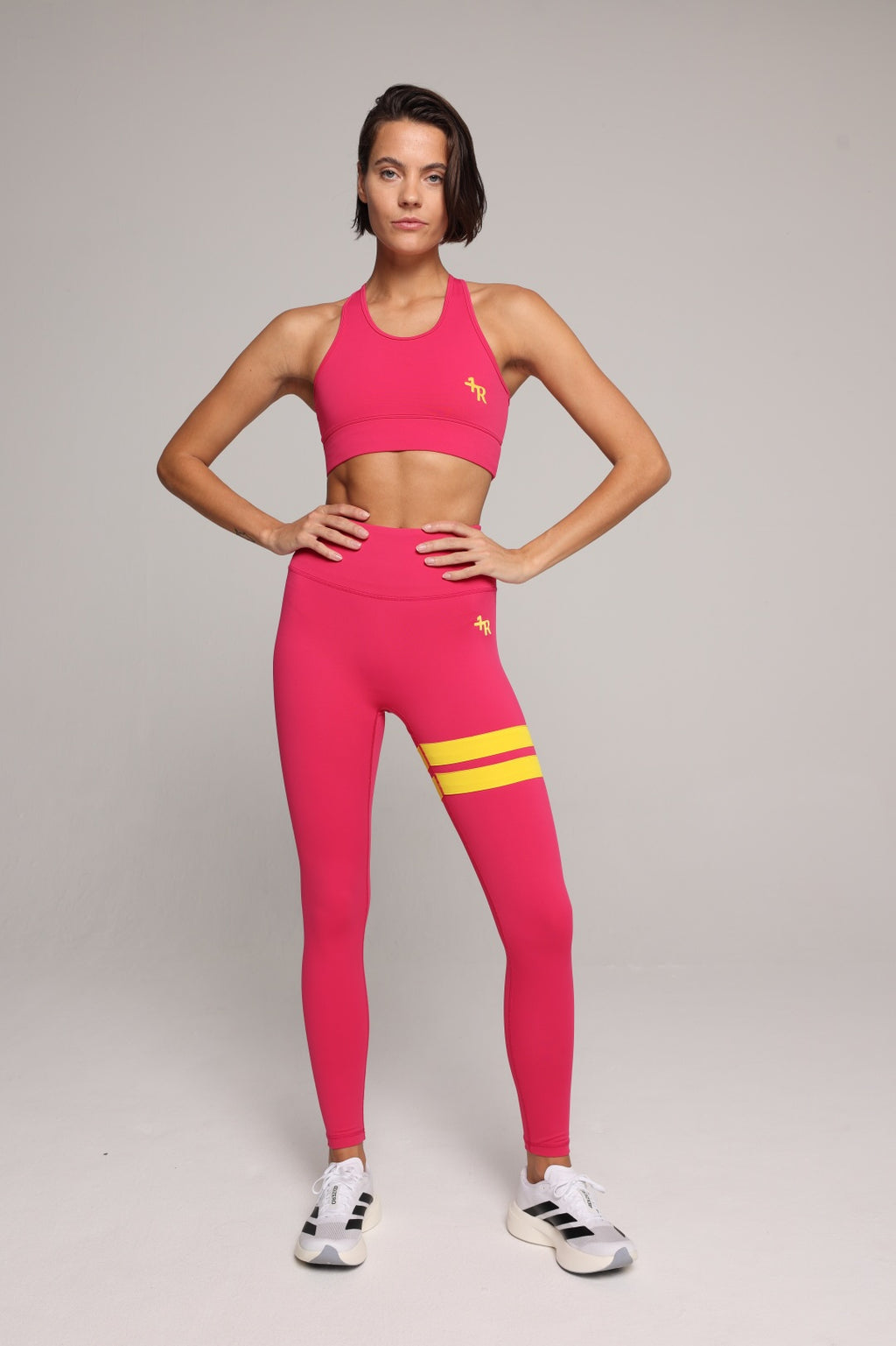 A woman modeling a bright pink athletic set consisting of a sports bra and high-waisted leggings with yellow stripes on the upper thigh, posed against a neutral light gray studio background. All by One More Reason, the European premium sportswear brand.