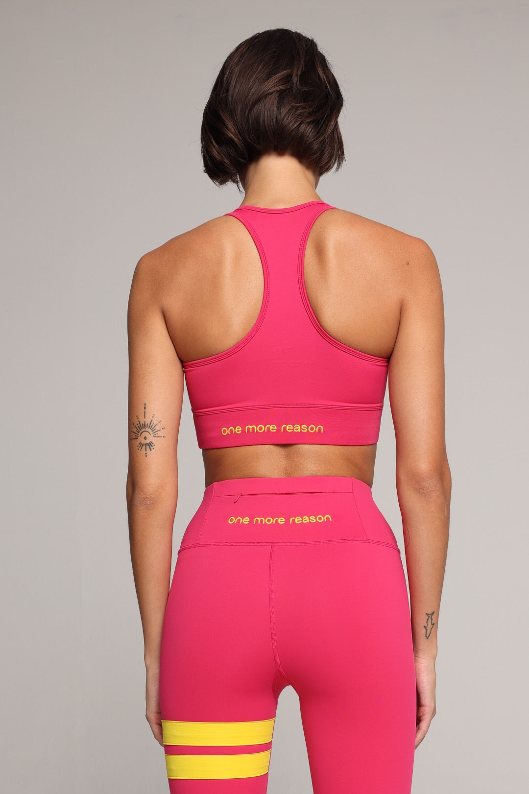 Rear view of a woman modeling a bright pink activewear set with yellow stripe accents on the leggings and yellow text reading 'one more reason' on the sports bra and waistband. All from One More Reason, the premium women's sportswear brand.