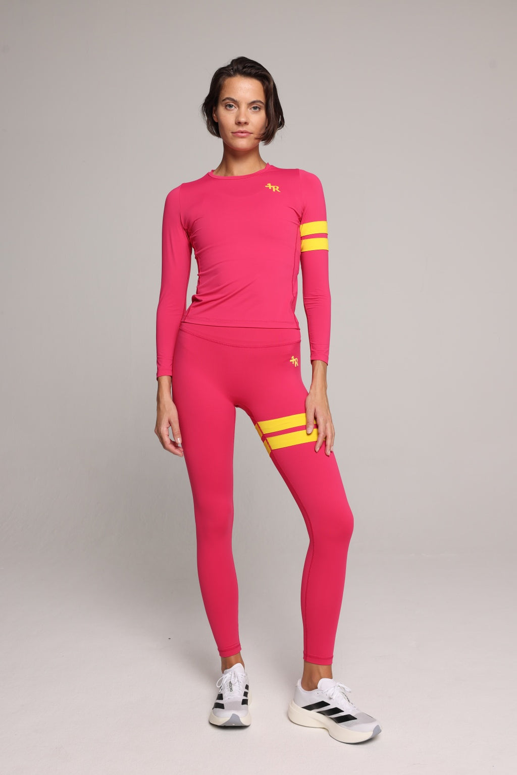 A woman in a bright pink long-sleeve athletic top and matching leggings set with yellow stripe accents on the sleeves and upper thigh, paired with white sneakers, against a neutral gray studio background. All by One More Reason, the European premium sportswear brand.
