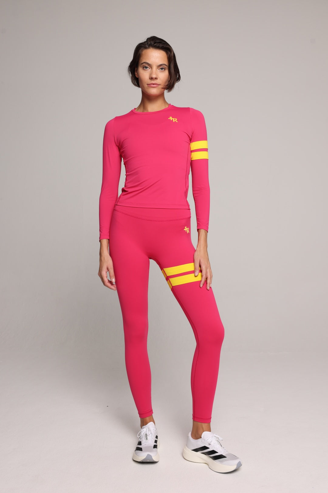 A woman in a bright pink long-sleeve athletic top and matching leggings set with yellow stripe accents on the sleeves and upper thigh, paired with white sneakers, against a neutral gray studio background. All by One More Reason, the European premium sportswear brand.