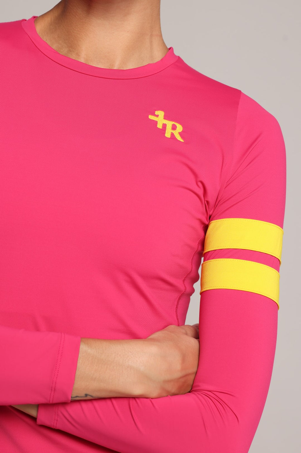 A close-up of a woman wearing a bright pink long-sleeve athletic top with a yellow embroidered logo on the left chest and two yellow stripes on the right sleeve, against a neutral gray background - by One More Reason, the European premium sportswear brand.