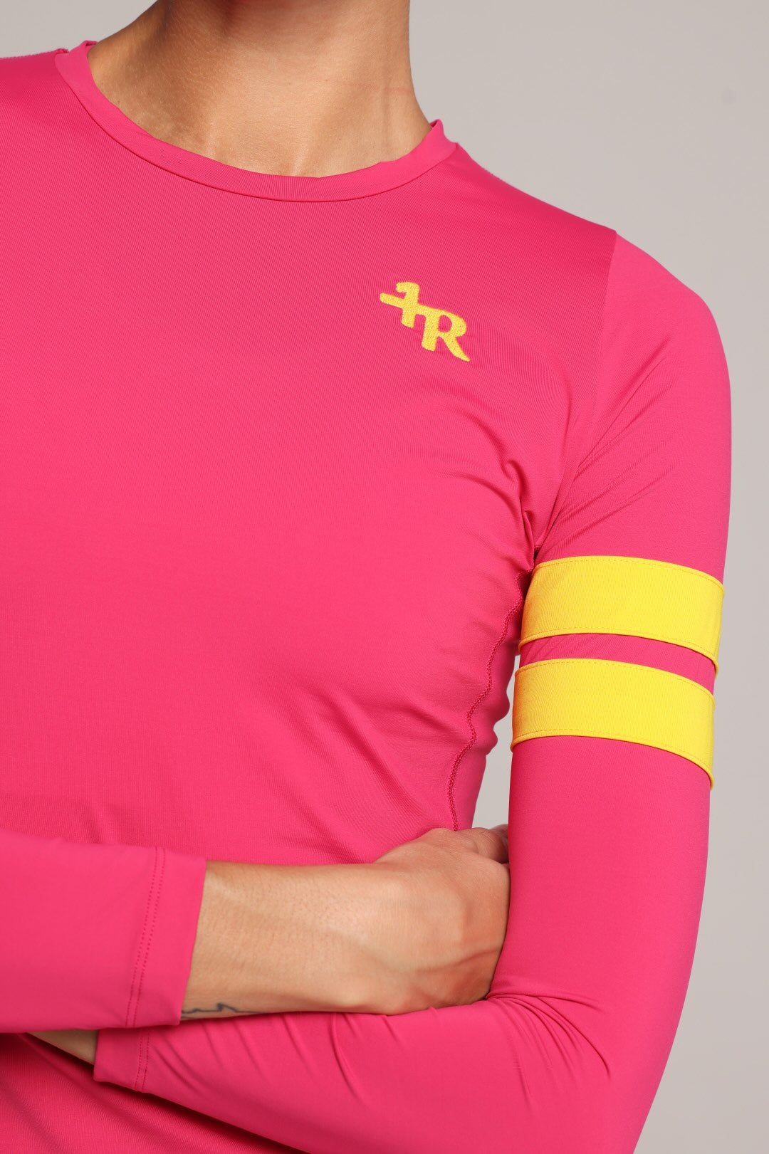 A close-up of a woman wearing a bright pink long-sleeve athletic top with a yellow embroidered logo on the left chest and two yellow stripes on the right sleeve, against a neutral gray background - by One More Reason, the European premium sportswear brand.