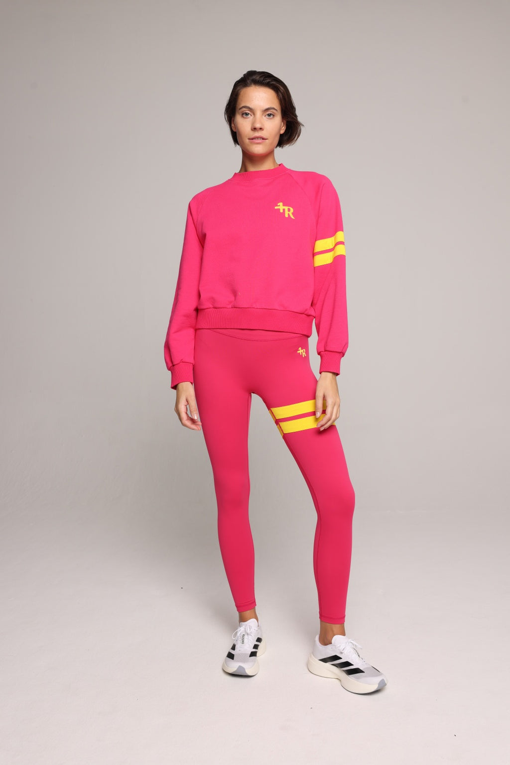 A woman wearing a bright pink sweatshirt and leggings with yellow stripes by One More Reason, the premium sportswear brand made in Europe in a studio setting 