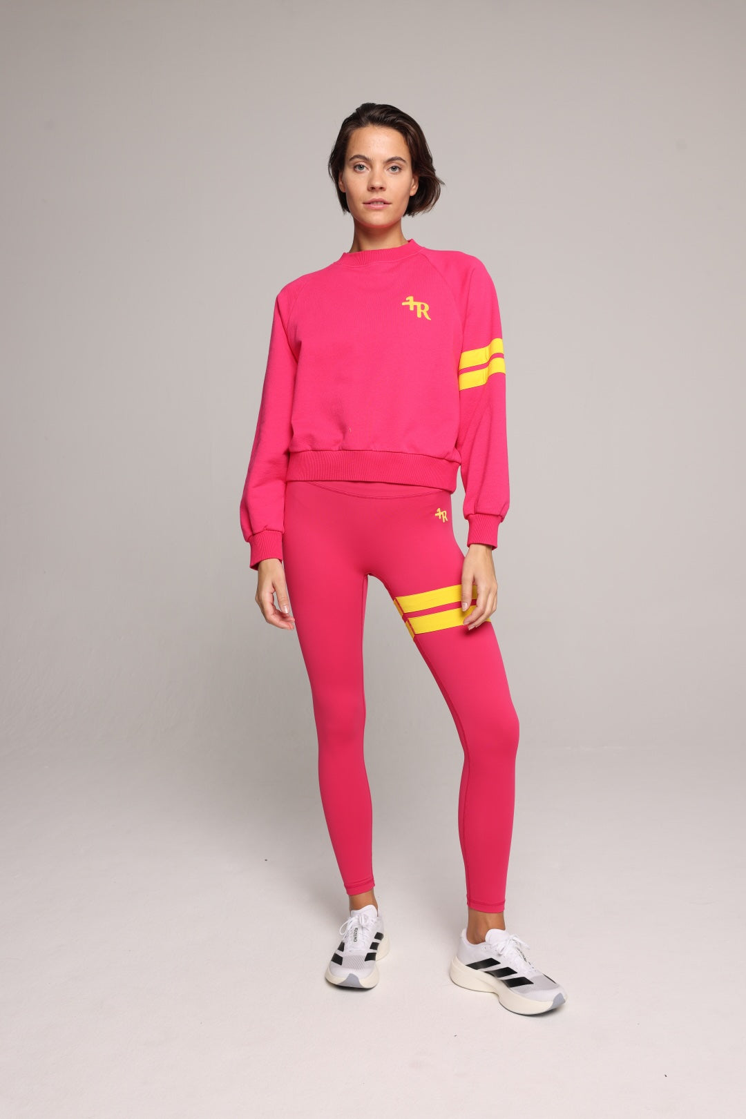 A woman wearing a bright pink sweatshirt and leggings with yellow stripes by One More Reason, the premium sportswear brand made in Europe in a studio setting 