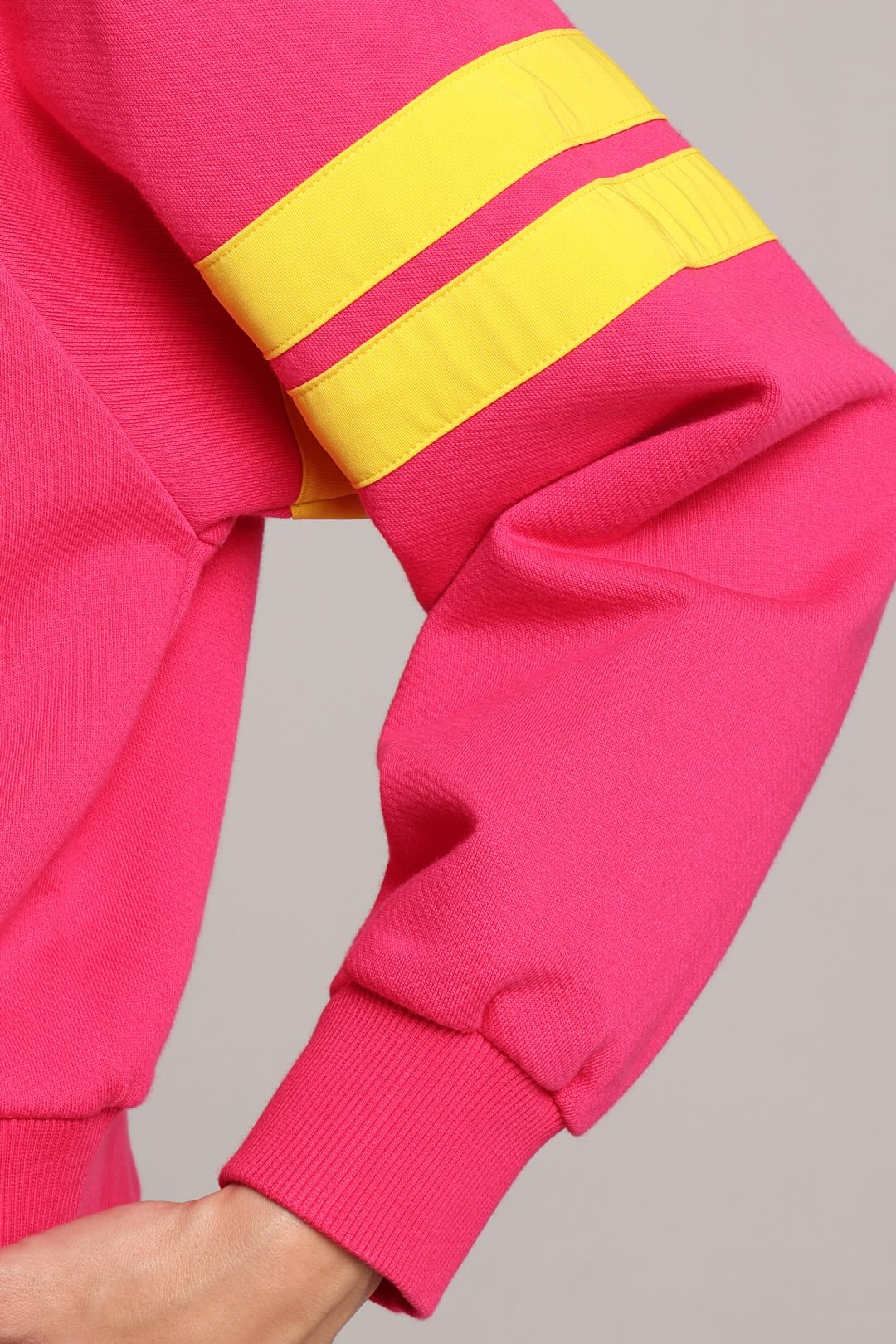 Close-up of a bright pink sweatshirt sleeve with two yellow stripes across the upper arm, shown against a plain light gray background. One More Reason premium sportswear.