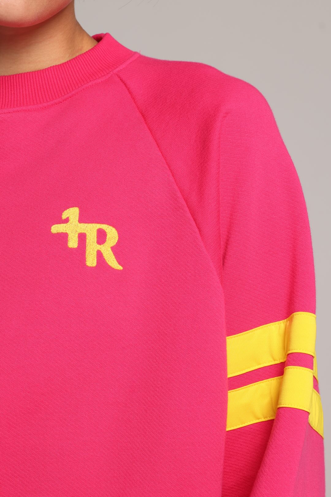A close-up of a bright pink crewneck sweatshirt with a yellow embroidered logo on the left chest and two yellow stripes on the right sleeve. One More Reason premium sportswear.
