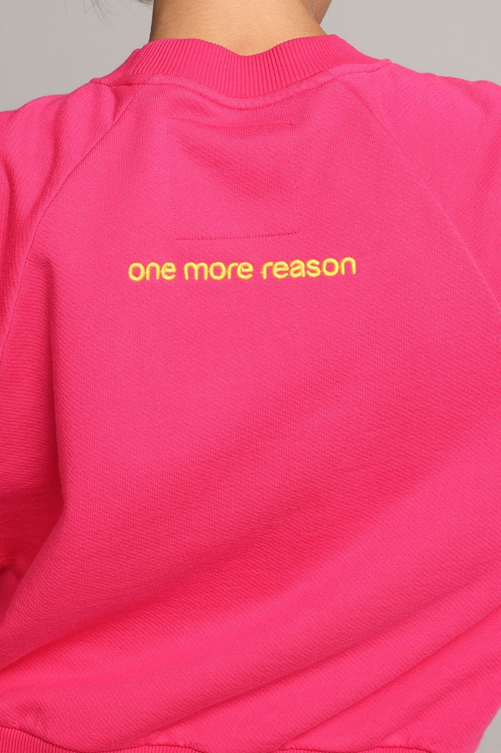 A close-up of a bright pink sweatshirt worn by a model, showing the back of the garment with yellow embroidered text reading 'one more reason'. One More Reason premium sportswear.