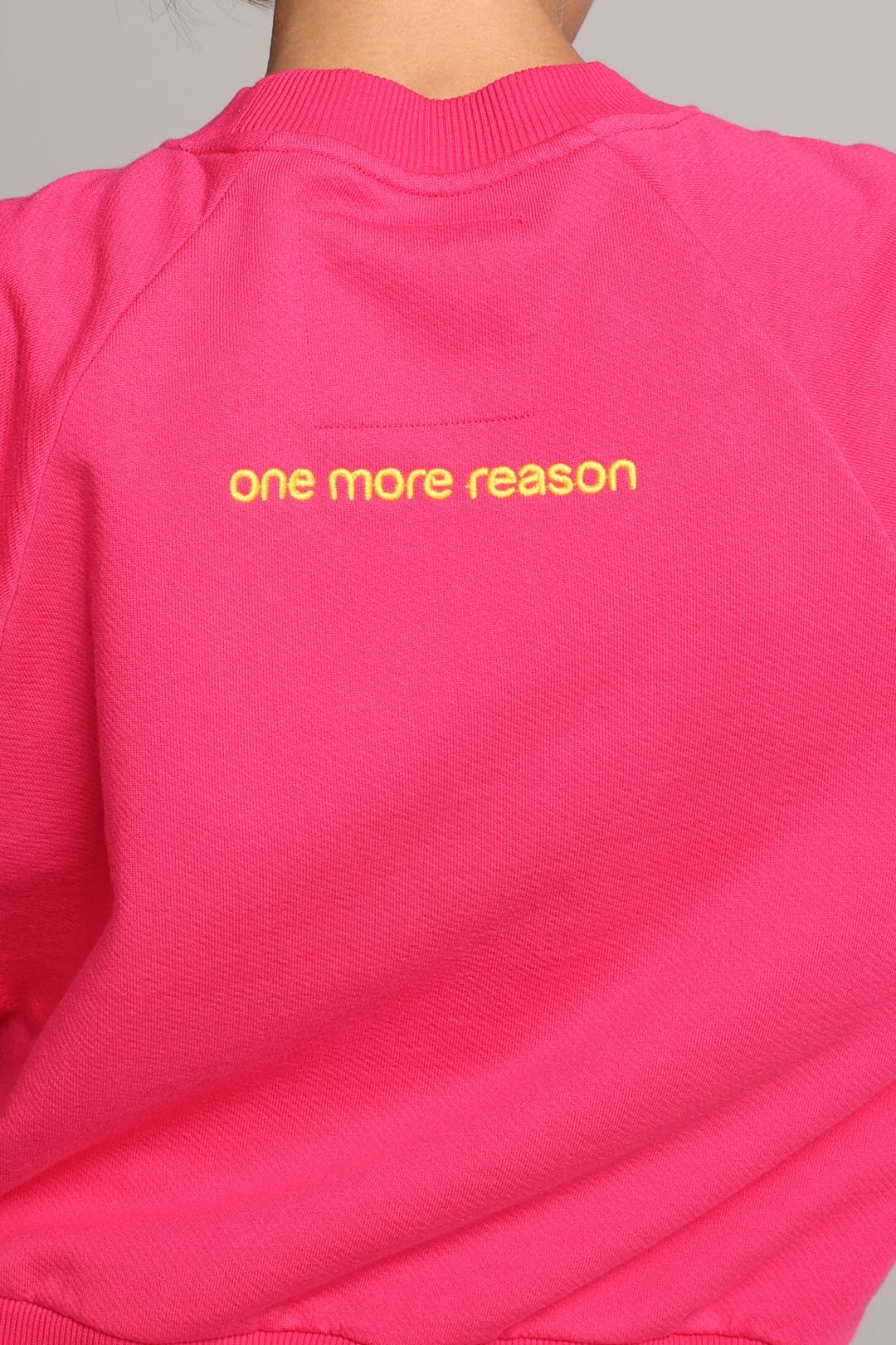 A close-up of a bright pink sweatshirt worn by a model, showing the back of the garment with yellow embroidered text reading 'one more reason'. One More Reason premium sportswear.