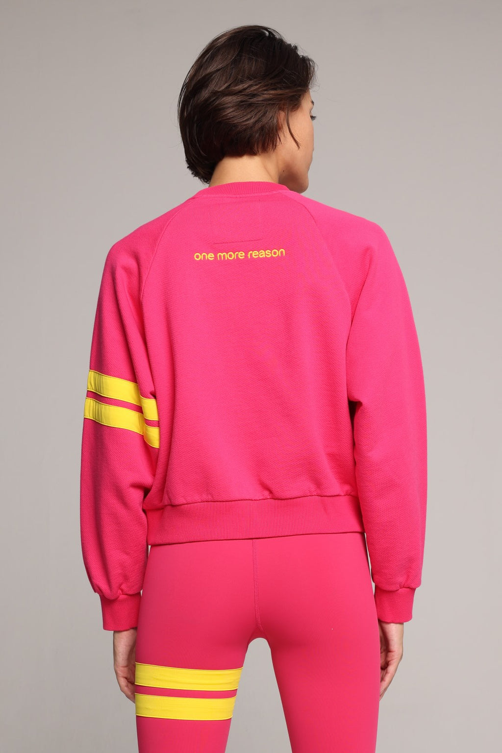 A woman showing from the back wearing a bright pink sweater and leggings with yellow stripes by One More Reason, the premium sportswear brand made in Europe in a studio setting 