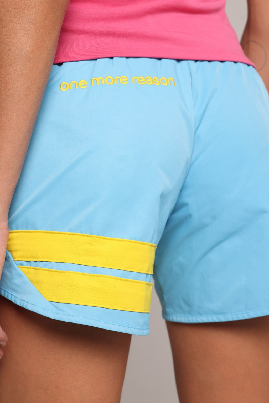 Close-up of a woman wearing light blue athletic shorts with a yellow stripe detail and yellow text reading 'one more reason' on the back pocket, paired with a pink top.