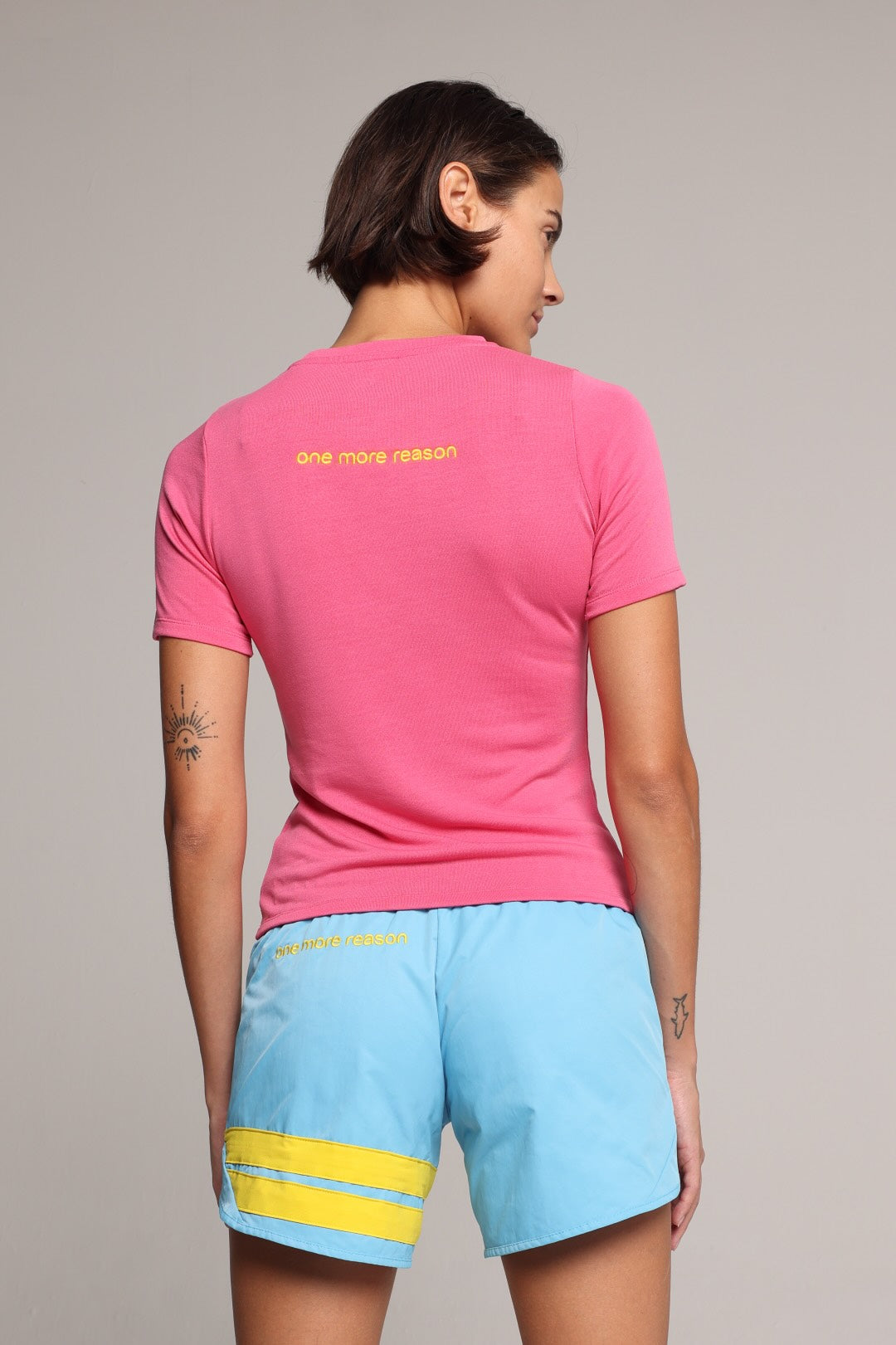 A woman shown from the back in a bright pink short-sleeve athletic t-shirt and light blue shorts with yellow stripes and logos, all by One More Reason, the premium European sportswear brand.