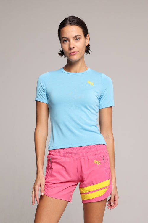 A woman in a light blue short-sleeve athletic t-shirt and bright pink athletic shorts with yellow accents, with a light gray studio background. All by One More Reason, the premium European sportswear brand.
