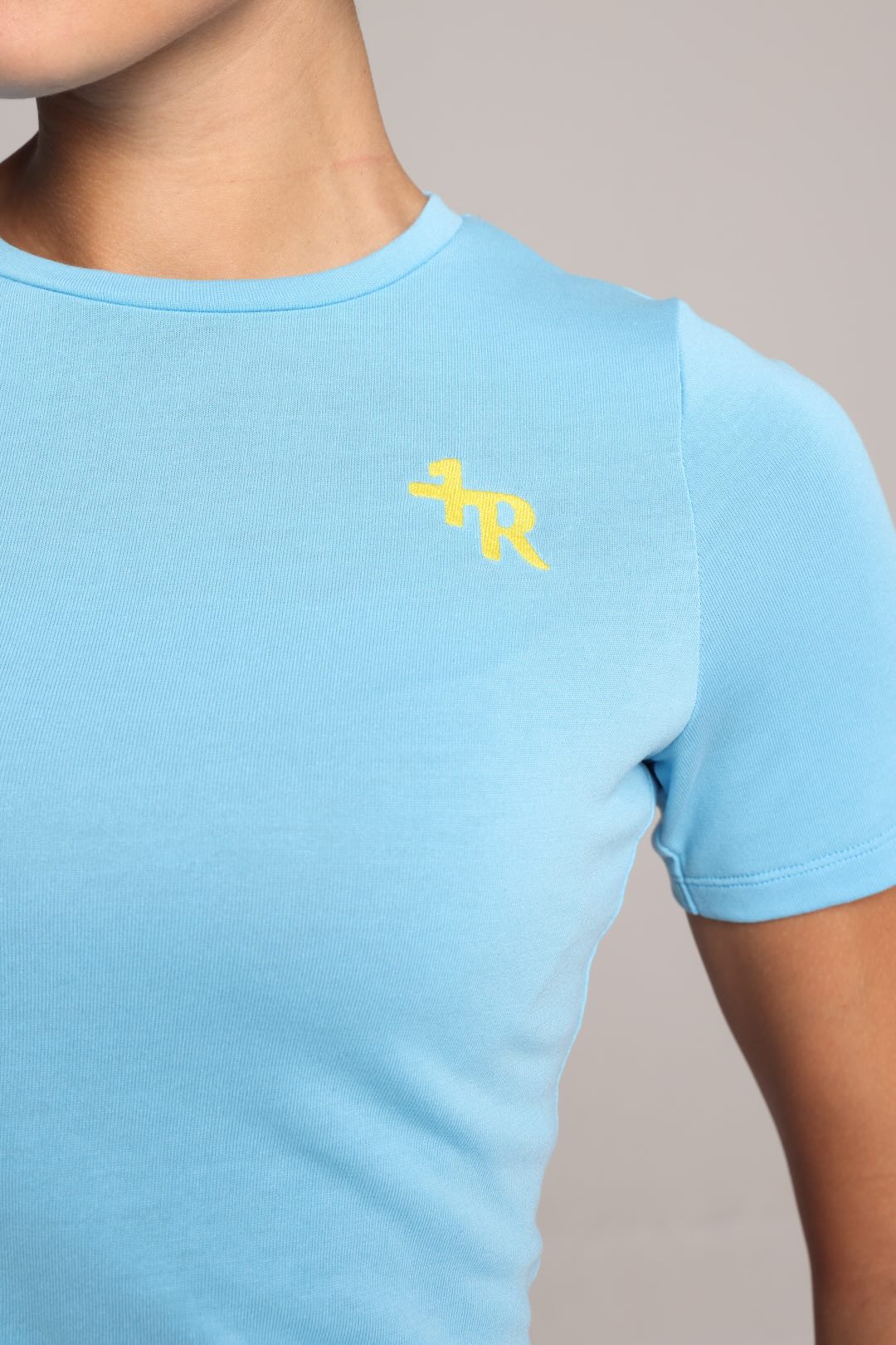 A close-up of a person wearing a light blue short-sleeve crewneck t-shirt with a small yellow embroidered logo on the left chest, set against a plain light gray background.