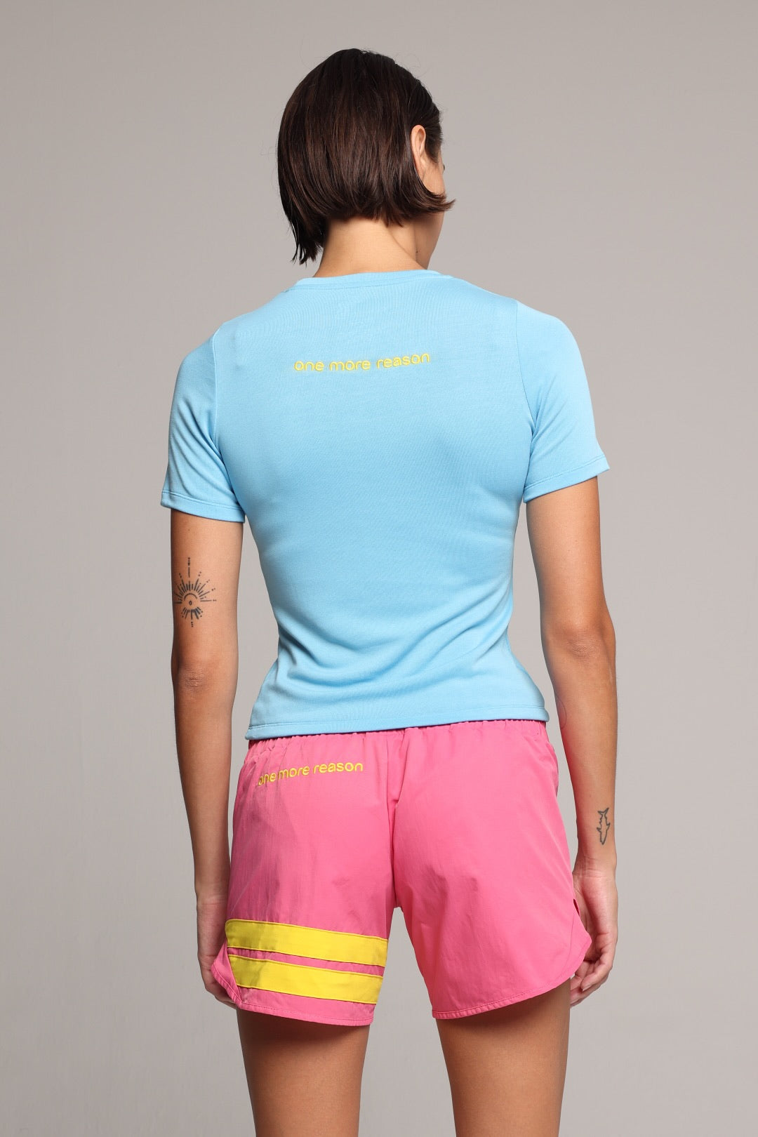 A woman shown from the back in a light blue short-sleeve athletic t-shirt and bright pink athletic shorts with yellow accents, with a light gray studio background. All by One More Reason, the premium European sportswear brand.