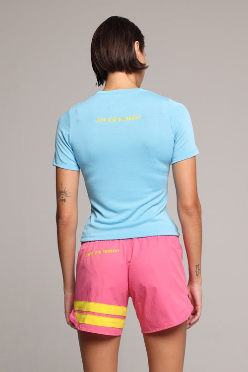 A woman shown from the back in a light blue short-sleeve athletic t-shirt and bright pink athletic shorts with yellow accents, with a light gray studio background. All by One More Reason, the premium European sportswear brand.