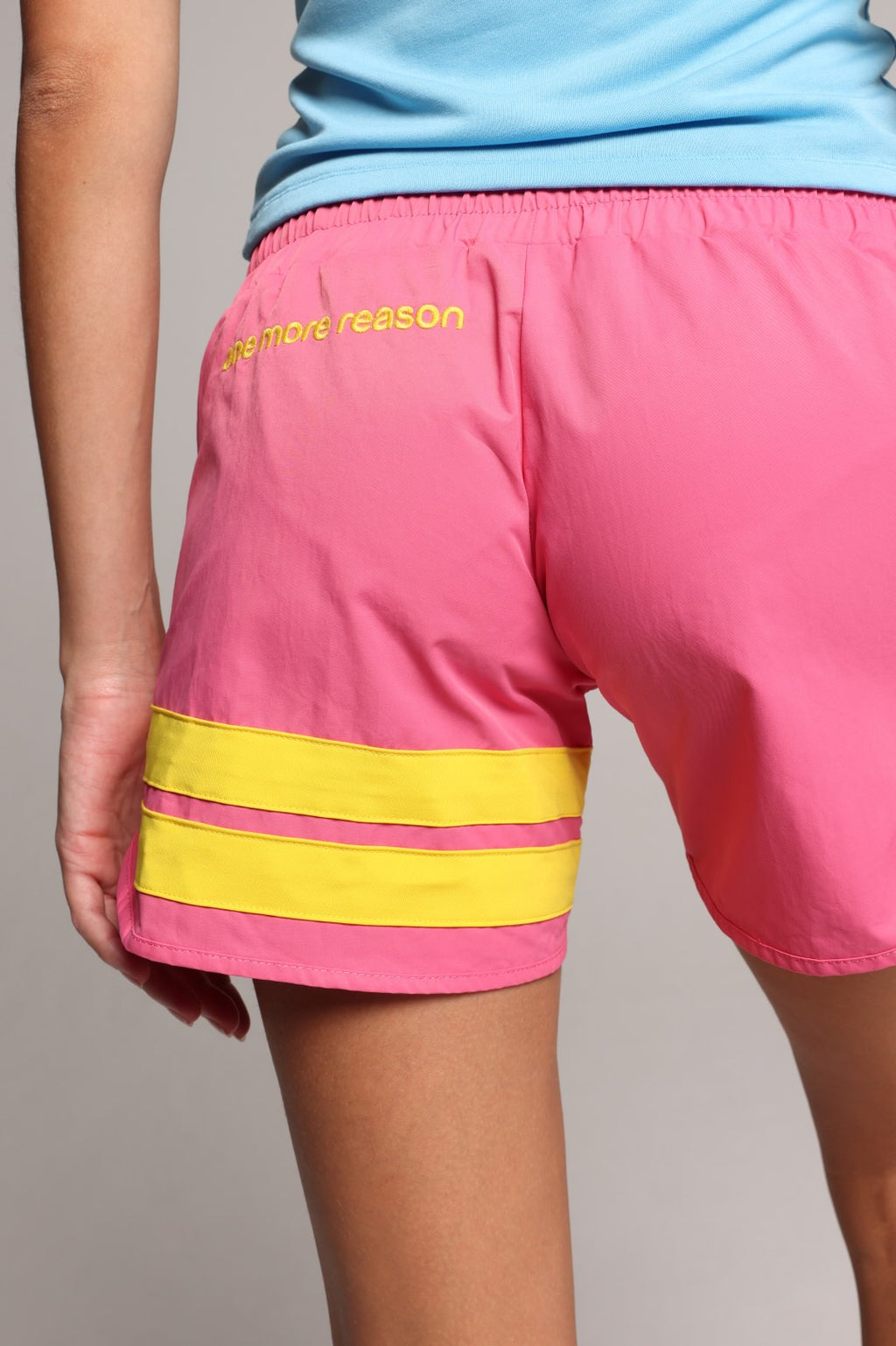 Pink athletic shorts with two horizontal yellow stripes near the hem and yellow text on the upper left leg, shown on a model wearing a light blue top against a neutral gray background.