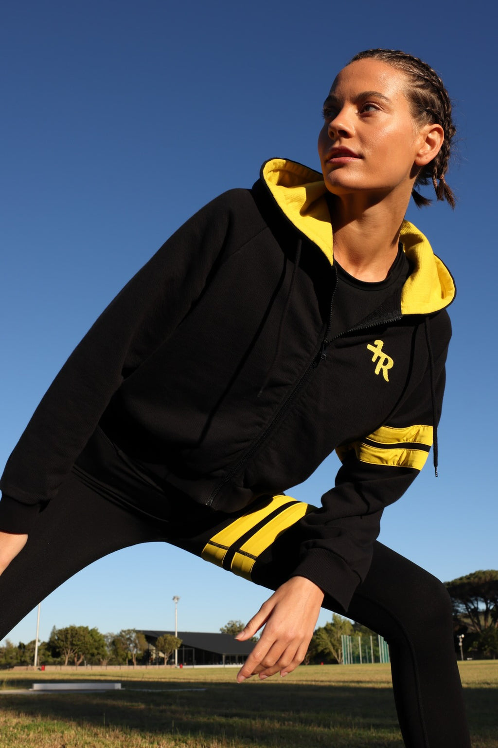 A woman wearing a black zip-up hoodie and matching black leggings with bright yellow accents, photographed outdoors against a clear blue sky. All from One More Reason, the European Premium Sportswear Brand.