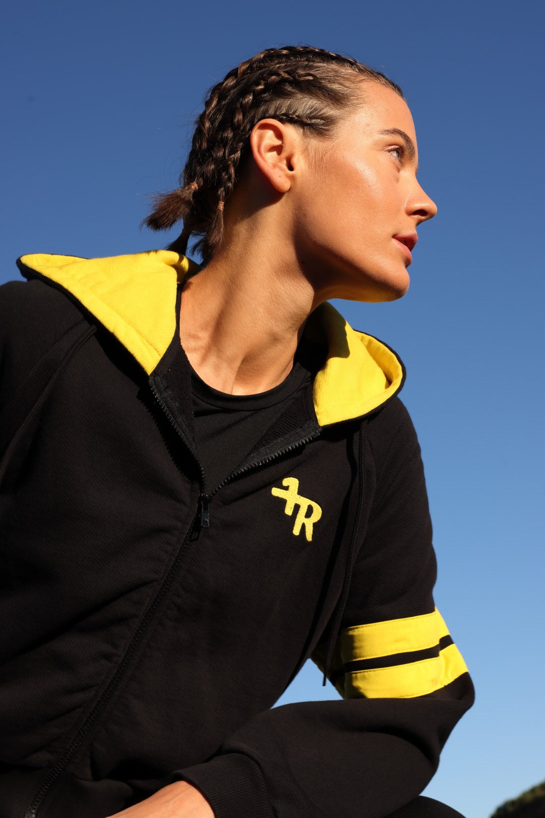 A woman in profile wearing a black zip-up hoodie with bright yellow hood lining and yellow stripe accents on the sleeve, photographed outdoors against a clear blue sky. All from One More Reason, the European Premium Sportswear Brand.