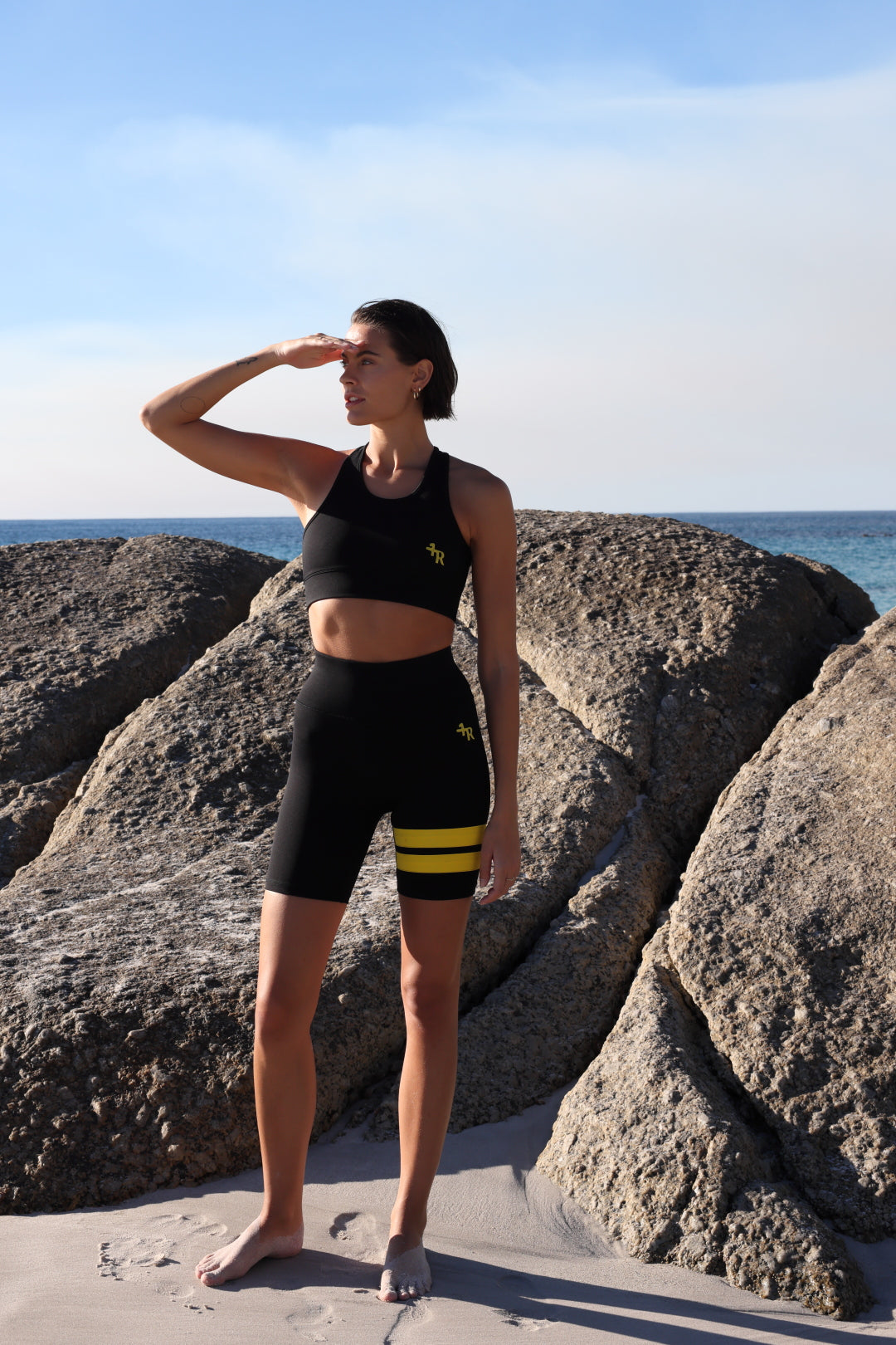 Woman in athletic black wear with yellow stripes by one more reason standing on rocks by the ocean