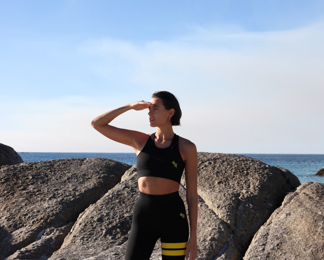 Woman in a premium black sports wear with yellow stripes by One More Reason is standing on rocks by the ocean
