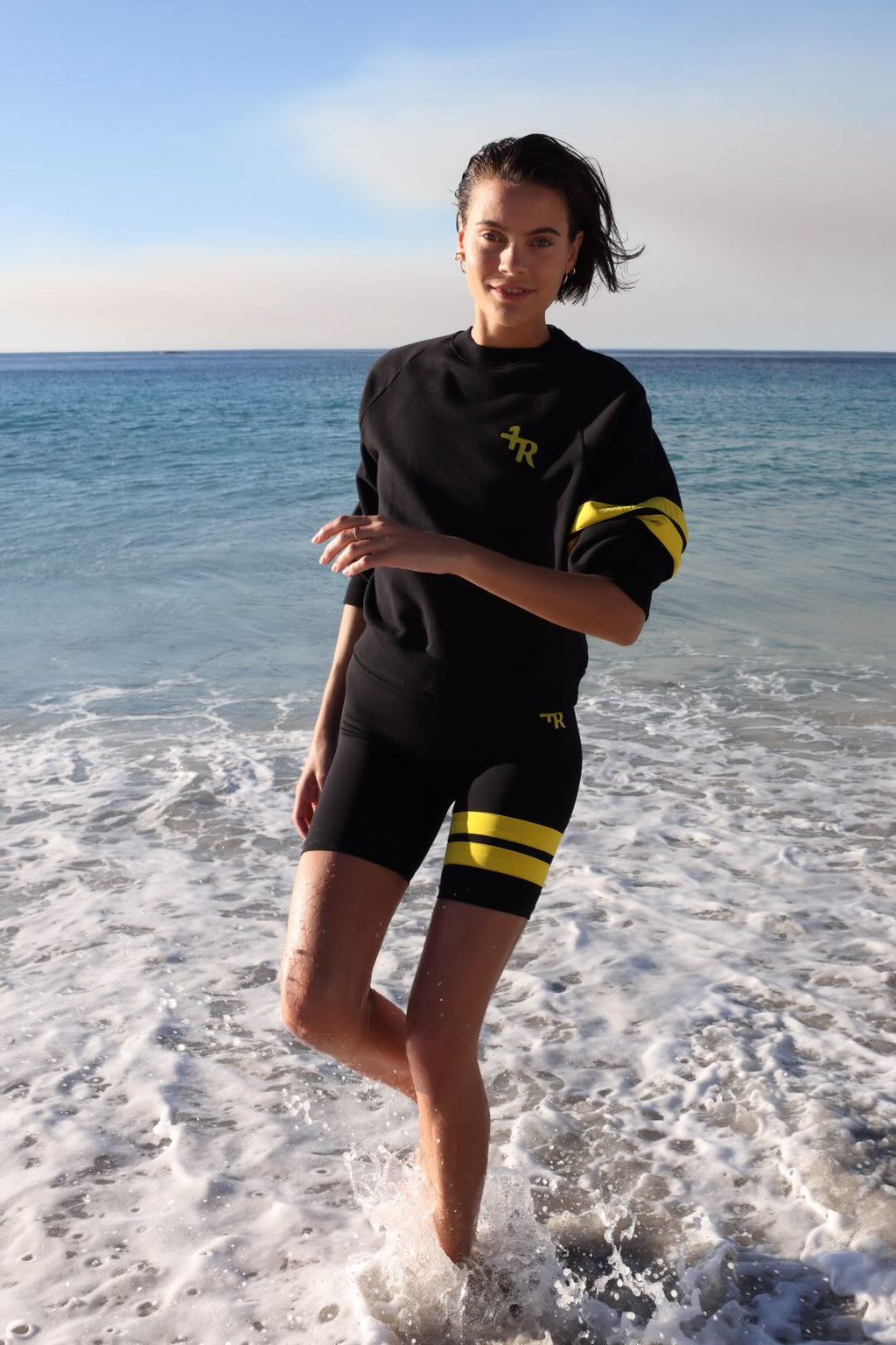 woman wearing a black biker shorts and a black sweater with yellow stripes by one more reason running in shallow water with a blue sky and ocean background