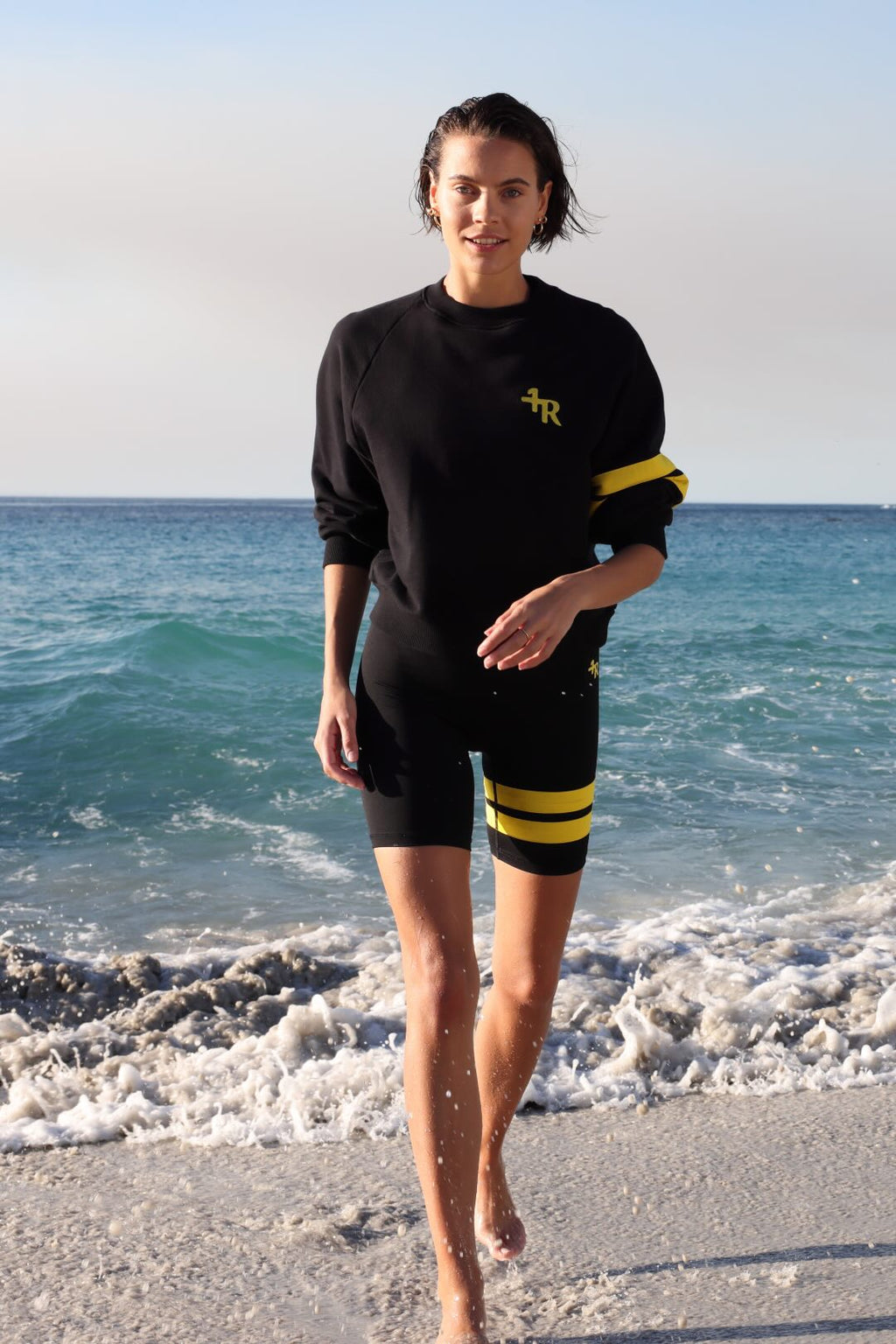 woman wearing a black short leggings and a black sweater with yellow stripes by one more reason standing in shallow water with a blue sky and ocean background