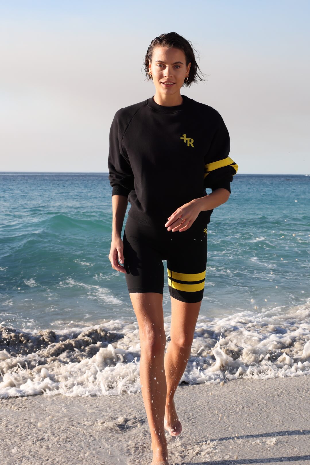 woman wearing a black short leggings and a black sweater with yellow stripes by one more reason standing in shallow water with a blue sky and ocean background
