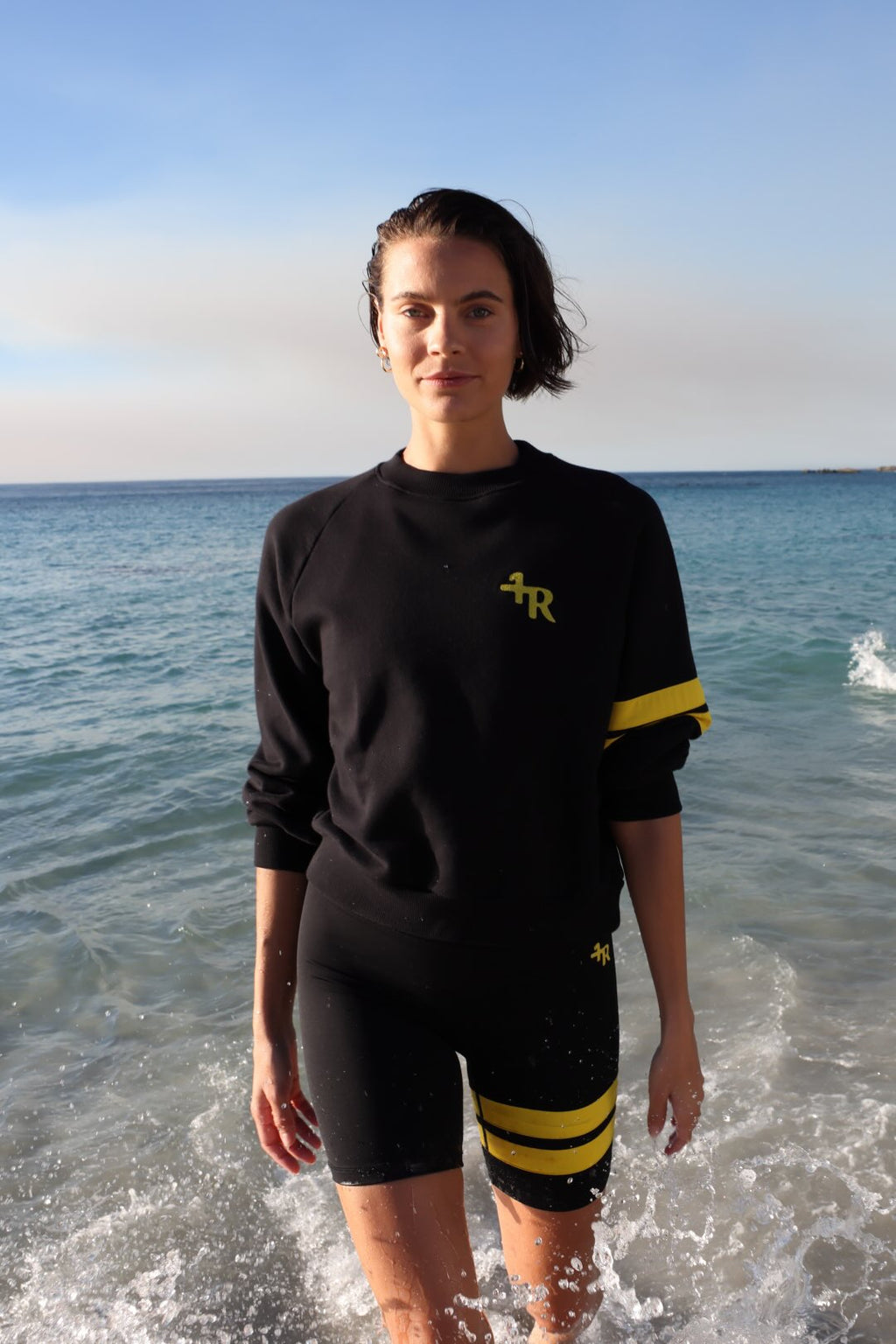 woman wearing a black short leggings and a black sweater with yellow stripes by one more reason standing in shallow water with a blue sky and ocean background