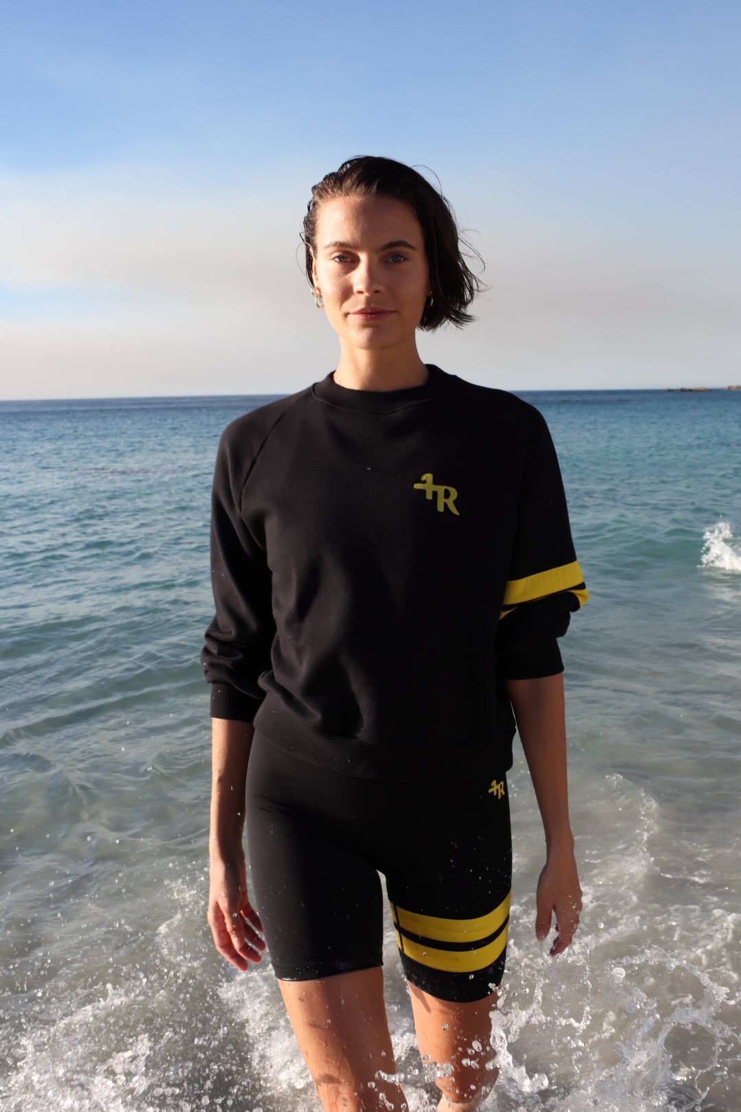 woman wearing a black short leggings and a black sweater with yellow stripes by one more reason standing in shallow water with a blue sky and ocean background