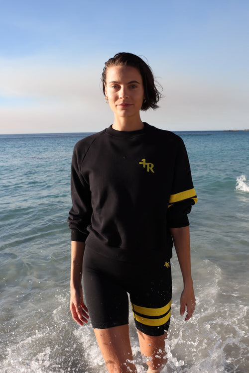 woman wearing a black short leggings and a black sweater with yellow stripes by one more reason standing in shallow water with a blue sky and ocean background