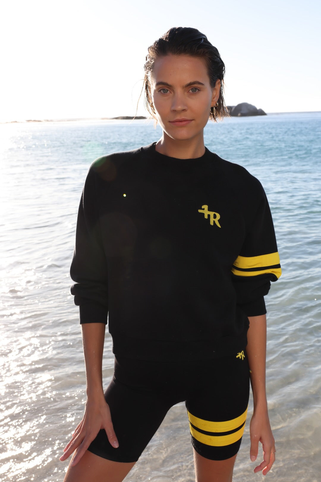 woman wearing a black short leggings and a black sweater with yellow stripes by one more reason standing in shallow water with a blue sky and ocean background