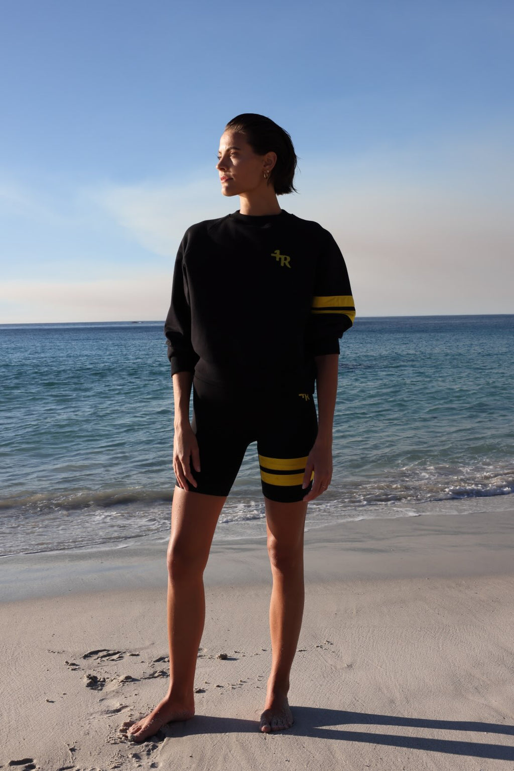 woman wearing a black biker shorts and a black sweater with yellow stripes by one more reason standing in shallow water with a blue sky and ocean background