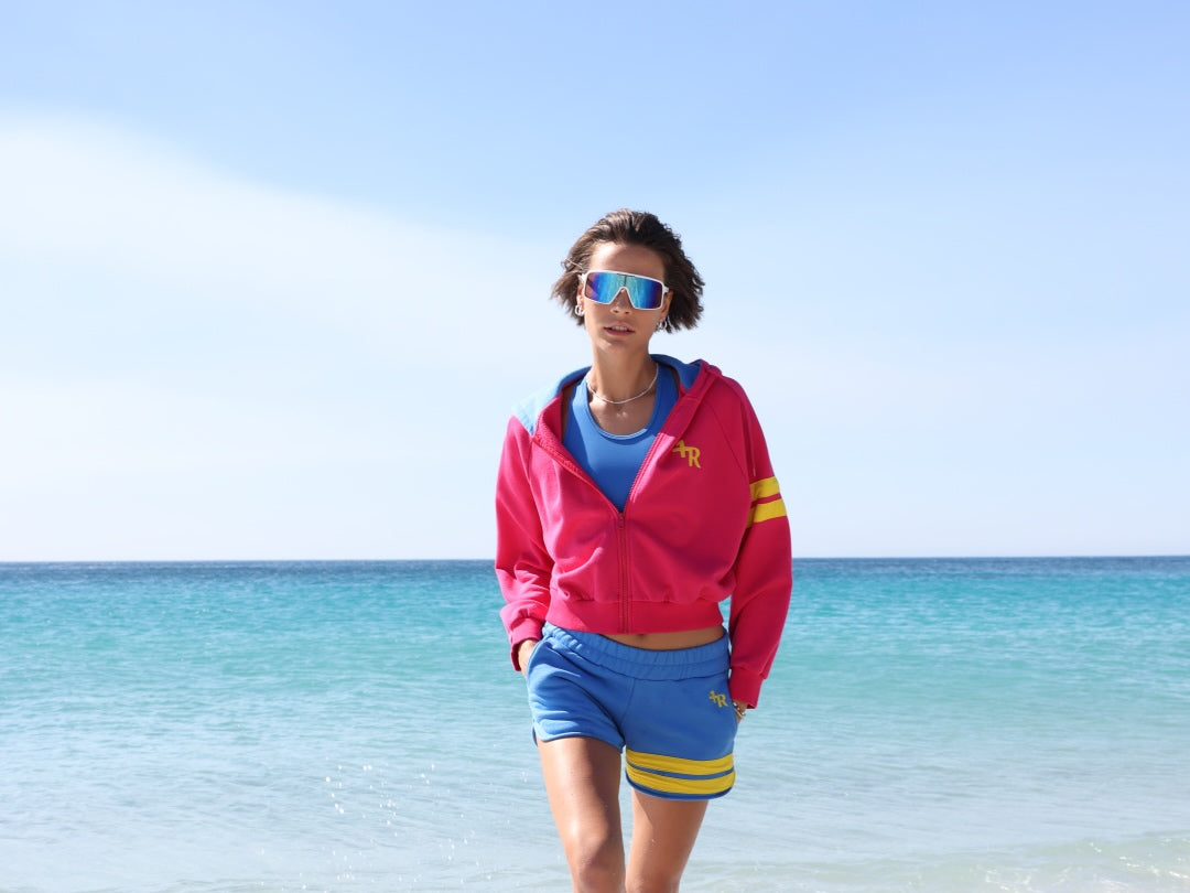 A woman stands on a sandy beach wearing a bright pink athletic crop top and matching shorts with yellow stripe accents, holding a blue jacket. All by One More Reason, the premium sportswear brand, made in Europe.