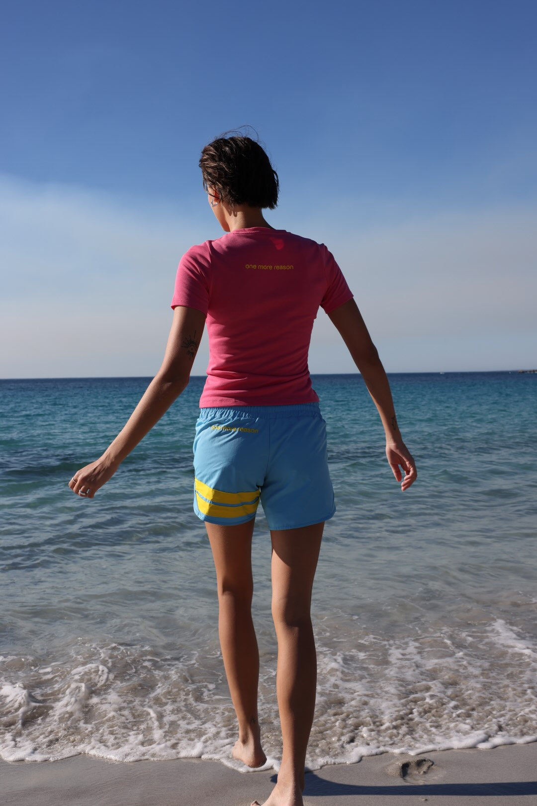 A woman shown from the back in a bright pink short-sleeve athletic t-shirt and light blue shorts with yellow stripes and logos running through shallow water at the beach. All by One More Reason, the premium European sportswear brand.
