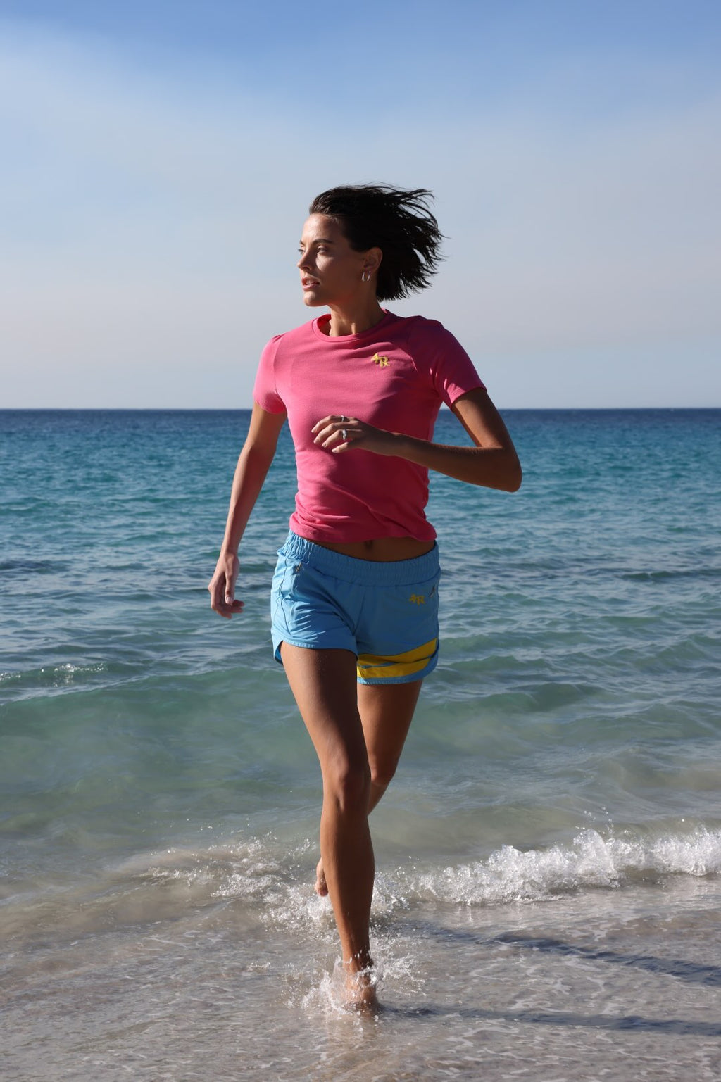 Woman running on  a beach in light blue performance shorts with yellow stripes and rose t-shirt by one more reason with ocean in the background