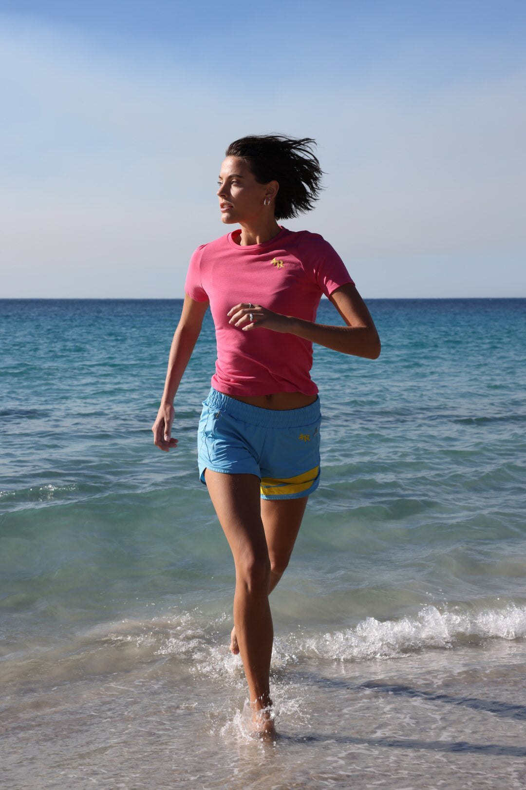 Woman running on  a beach in light blue performance shorts with yellow stripes and rose t-shirt by one more reason with ocean in the background
