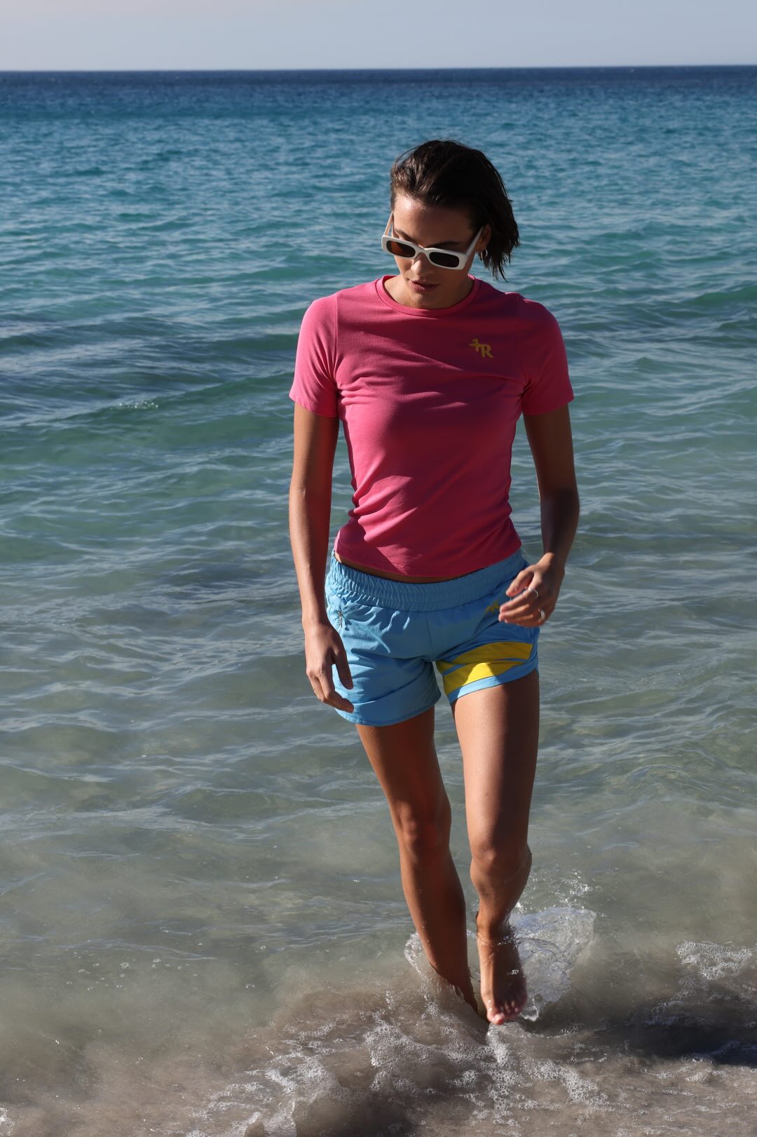 Woman in premium pink t-shirt and blue performance shorts with yellow stripe by one more reason running on a beach