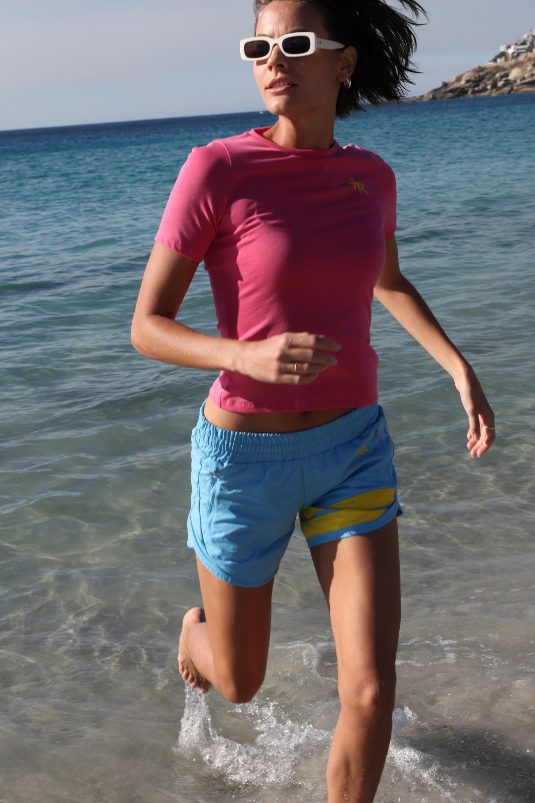 A woman in a bright pink short-sleeve athletic t-shirt and light blue shorts with yellow stripes and logos running through shallow water at the beach. All by One More Reason, the premium European sportswear brand.