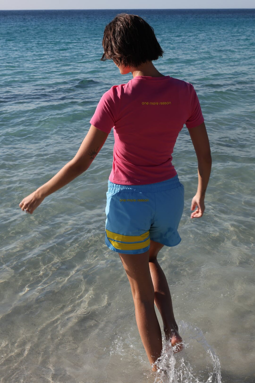 Woman in premium pink t-shirt and blue performance shorts with yellow stripe by one more reason running on a beach
