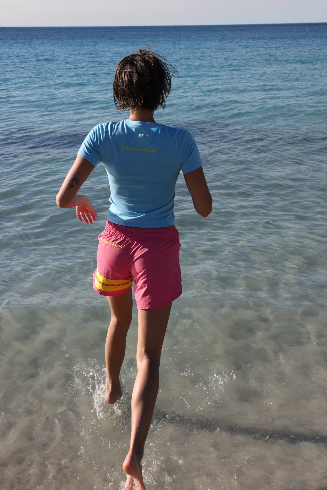 A woman seen from behind running at the beach in  a light blue short-sleeve athletic t-shirt and bright pink athletic shorts with yellow accents. All by One More Reason, the premium European sportswear brand. 