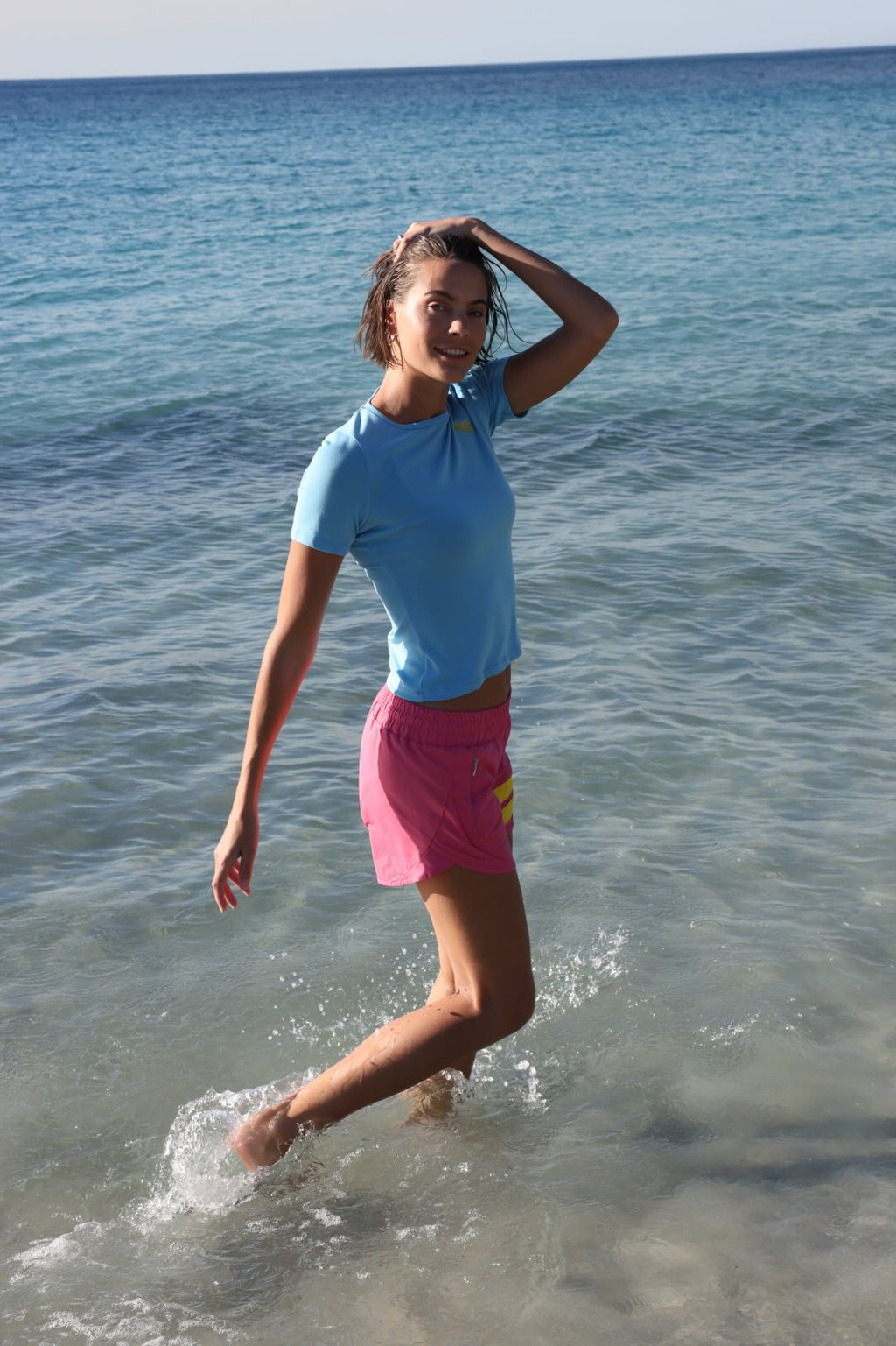 Woman running on a beach with ocean in the background wearing rose performance shorts with yellow stripes and a blue t-shirt by one more reason