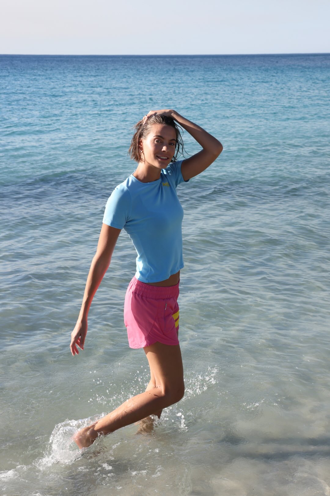 A woman walking through shallow ocean water wearing a light blue short-sleeve swim top and bright pink swim shorts.