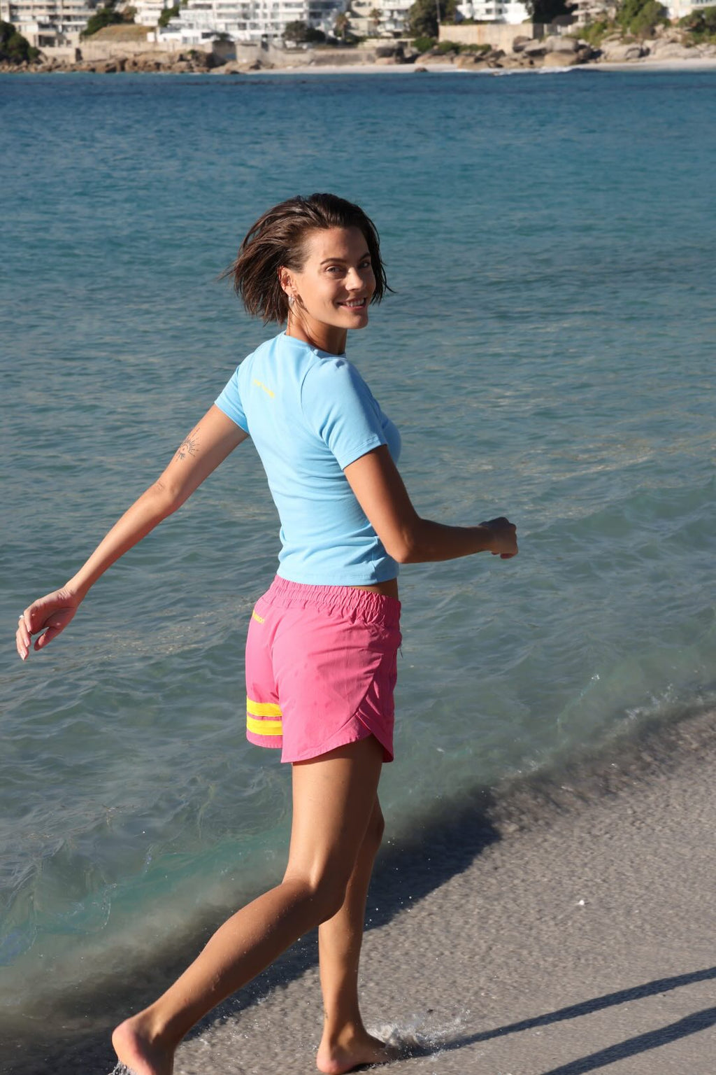 Woman running on a beach with ocean in the background wearing rose performance shorts with yellow stripes and a blue t-shirt by one more reason