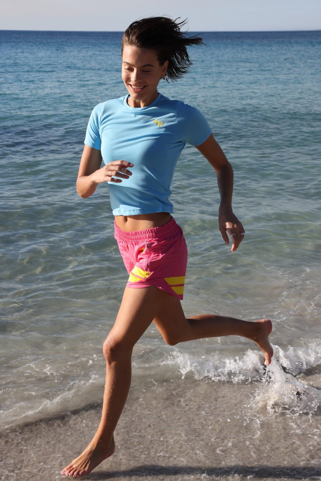 Woman running on a beach with ocean in the background wearing rose performance shorts with yellow stripes and a blue t-shirt by one more reason