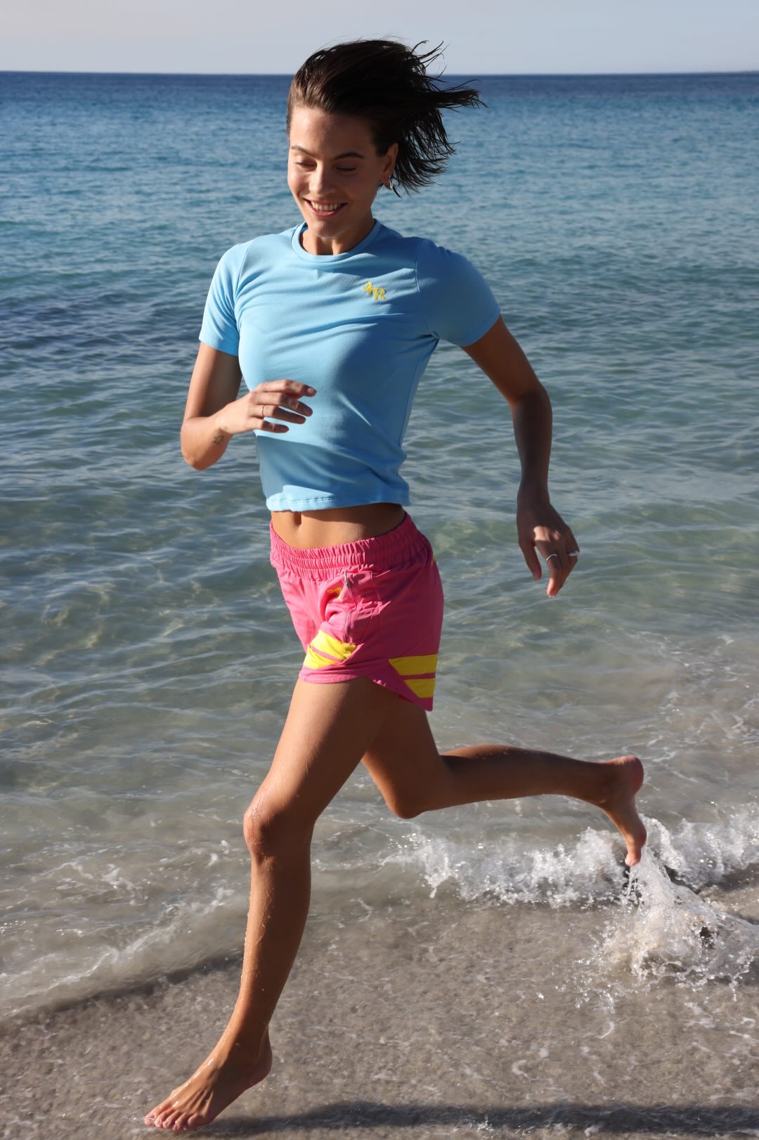 Woman running on a beach with ocean in the background wearing rose performance shorts with yellow stripes and a blue t-shirt by one more reason