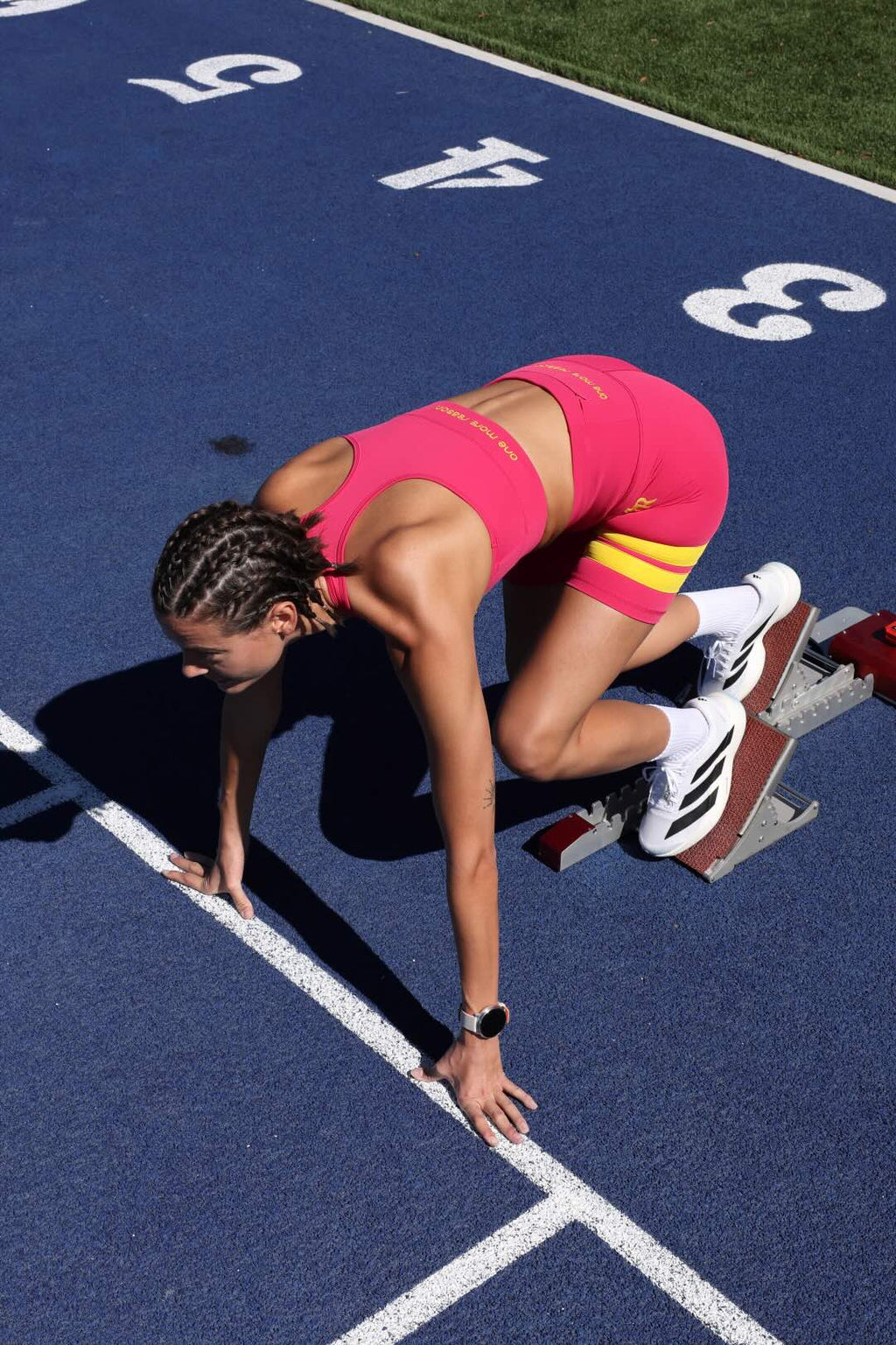 A female athlete in a pink athletic outfit with yellow stripes crouches in a starting position on a blue running track. All by One More Reason, European's premium sportswear brand.