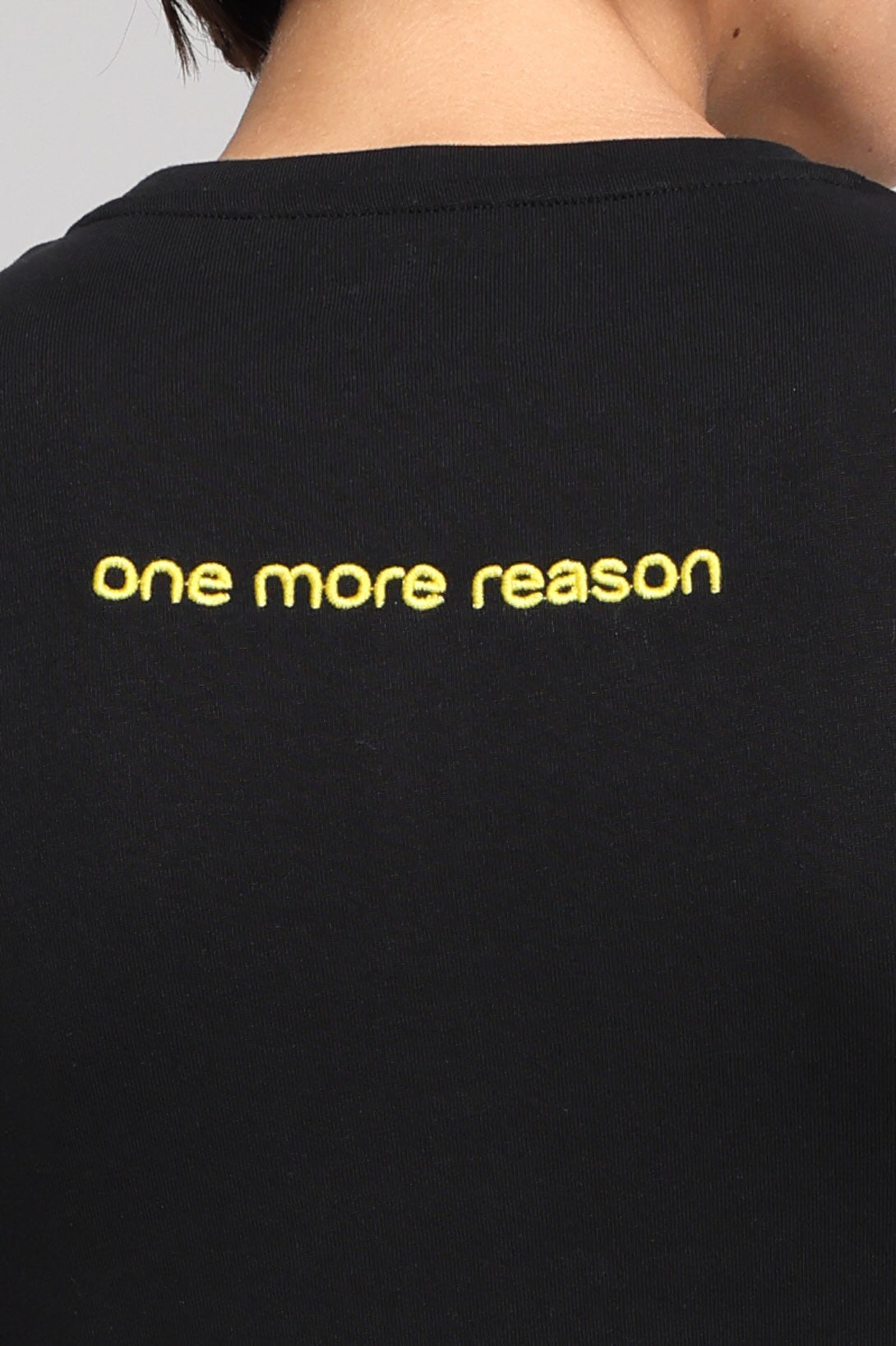 Close-up of the back of a person wearing a black T-shirt with the phrase 'one more reason' embroidered in yellow across the upper back.
